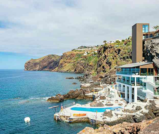 Utsikt over kystlandskap med hotellbygning og utendørsbasseng på en klippe ved havet. (AI-generert tekst)