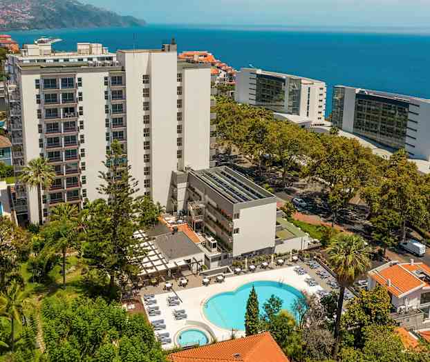 Hotellbygning med basseng og omkringliggende beplantning, sjø i bakgrunnen. (AI-generert tekst)