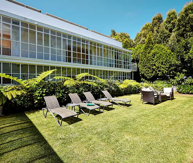 Bilde av en hage med solsenger, sittegruppe og grønne planter foran en bygning med store vinduer. (AI-generert tekst)