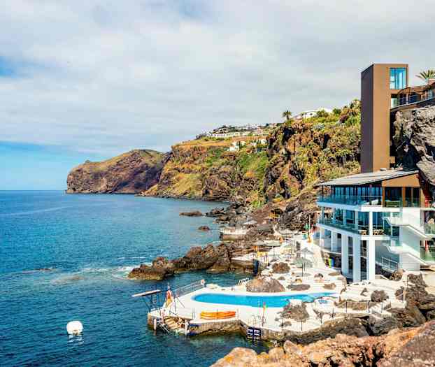 Kystlandskap med en moderne bygning og bassengområde ved klippene, omgitt av havet og frodige skråninger. (AI-generert tekst)