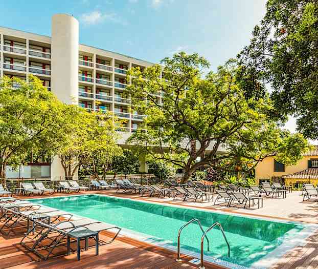 Utendørsbasseng med solsenger omgitt av trær og bygninger. (AI-generert tekst)