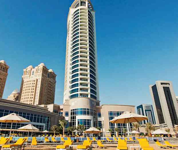 Bildet viser en moderne høy bygning omgitt av flere andre bygninger og et område med gule solsenger og parasoller. (AI-generert tekst)