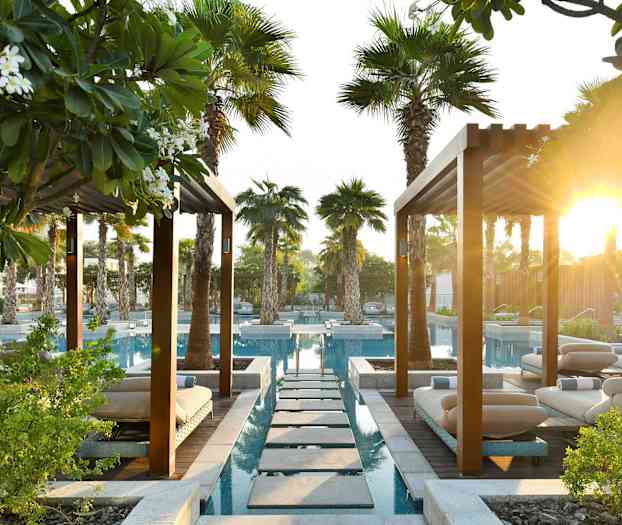 En hageanlegg med bassenger, solsenger og palmer under pergolatak med solskinn i bakgrunnen. (AI-generert tekst)