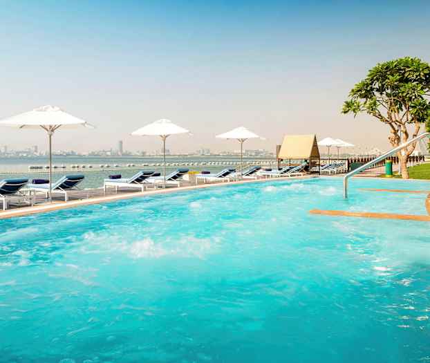 Utendørsbasseng med solsenger og parasoller ved sjøen, bysilhuetten er synlig i bakgrunnen. (AI-generert tekst)