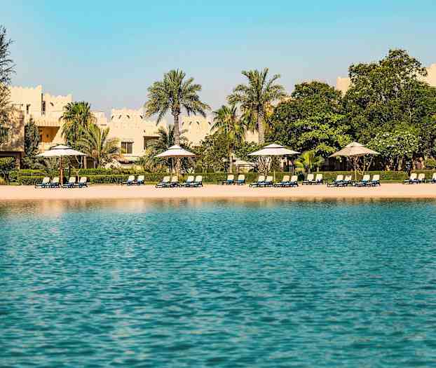 Utsikt over en strand med solsenger og parasoller, omgitt av palmer og bygninger med tradisjonell arkitektur i bakgrunnen. (AI-generert tekst)