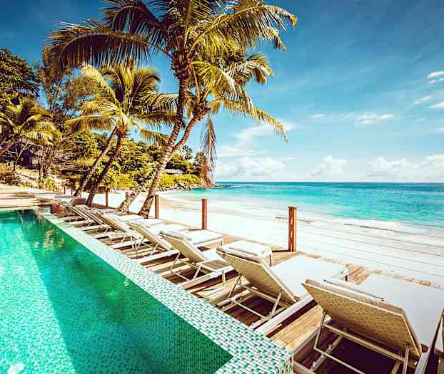 Utendørsbasseng med solsenger ved en sandstrand med klart blått vann. (AI-generert tekst)