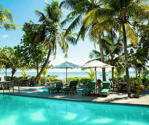 Et bilde av et basseng omgitt av solsenger med parasoller, omgitt av palmer og grøntområder med utsikt over havet. (AI-generert tekst)