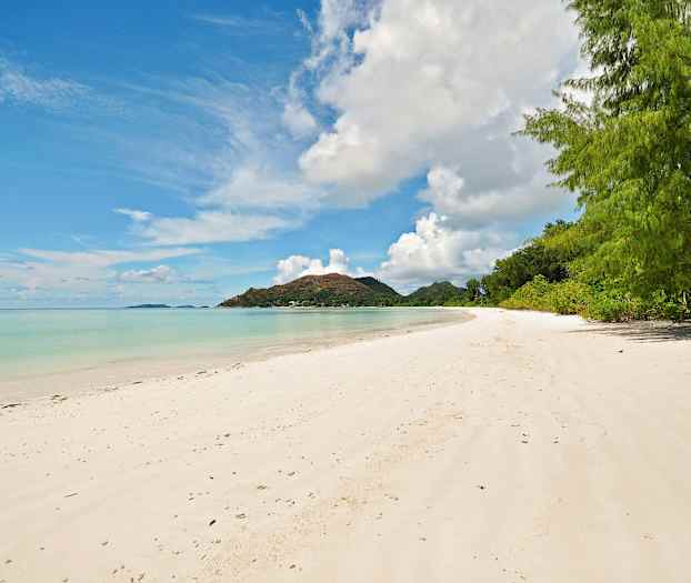 En sandstrand med klart vann og grønn vegetasjon i bakgrunnen. En fjelltopp kan ses på horisonten. (AI-generert tekst)