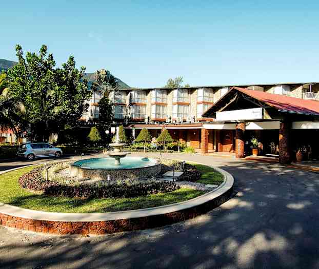 Hotellkompleks med store vinduer, velstelt hage og en fontene, plassert mot en bakgrunn av fjell. (AI-generert tekst)