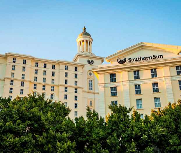 En lys bygning med flere etasjer og et tårn er synlig bak frodig vegetasjon. (AI-generert tekst)