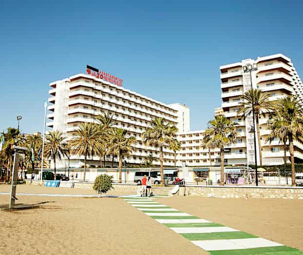 En strandpromenade foran høye leilighetsbygninger med palmetrær i forgrunnen. (AI-generert tekst)