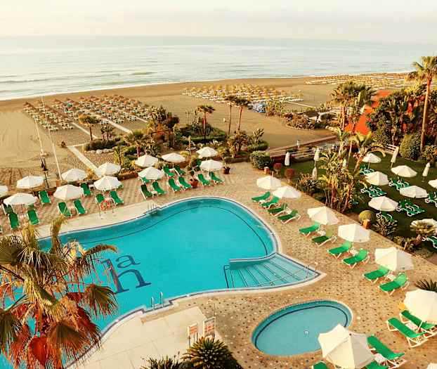 Et utendørs basseng med solsenger og parasoller rundt, strand synlig i bakgrunnen. (AI-generert tekst)