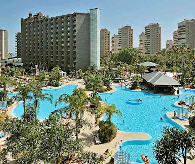 Et hotellområde med stort basseng og solsenger, omgitt av palmer og høyhus. (AI-generert tekst)