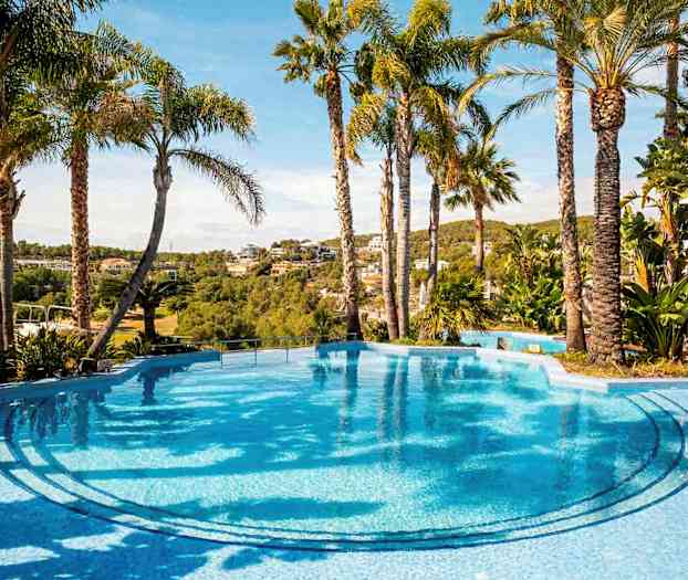 Et klart blått basseng omgitt av palmer med utsikt over et frodig landskap og hus i bakgrunnen. (AI-generert tekst)