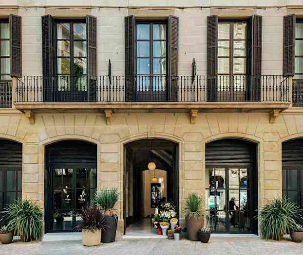 Bilde av fasaden til en bygning med flere høye vinduer og en inngangspart med dekorasjoner av blomsterved. (AI-generert tekst)