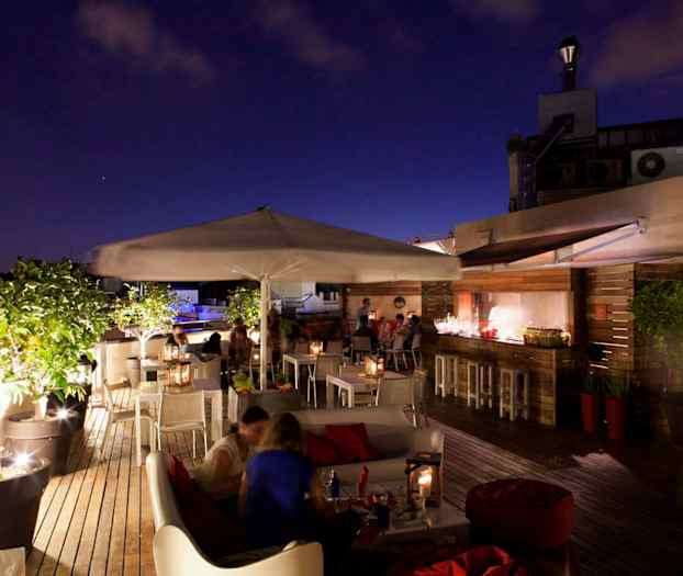 En takterrasse med sittegrupper, bord, parasoller og grønt, opplyst i kveldsomgivelser. (AI-generert tekst)