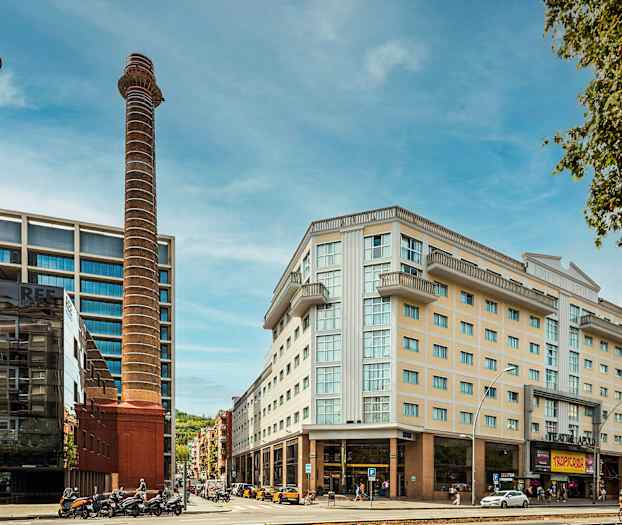 Bilde av et bymiljø med moderne bygninger, to høye skorsteiner og noen mennesker og kjøretøy i forgrunnen. (AI-generert tekst)