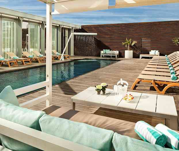 Et utendørs svømmebasseng med solsenger og håndklær på et tregulv, samt et bord med dekorasjoner og parasoll. (AI-generert tekst)