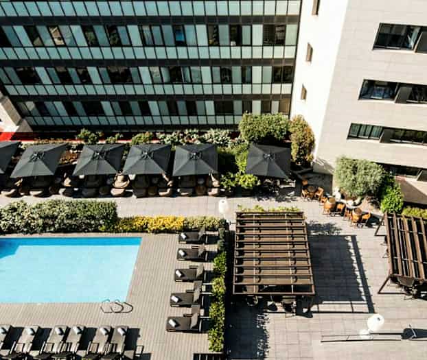 Flyfoto av et basseng med solsenger, parasoller og omgitt av bygninger. (AI-generert tekst)