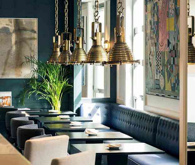 Interiør av en restaurant med mørkeblå vegger, kunstverk, hengende lamper og sitteplasser ved bord. (AI-generert tekst)