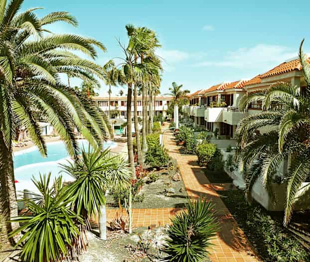 Bilde av et ferieanlegg med palmer, hager og et basseng. Bygninger med terrakottafargede tak er synlige i bakgrunnen. (AI-generert tekst)