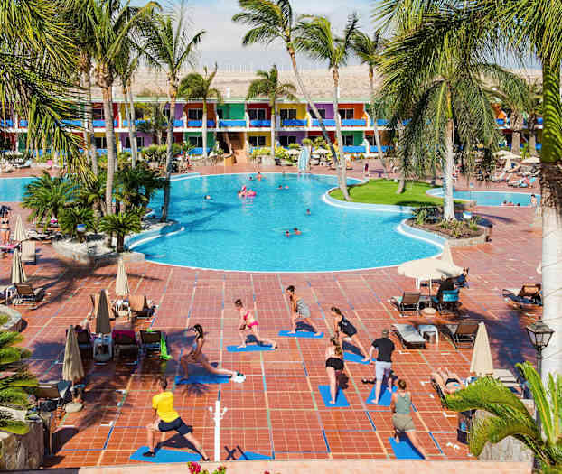 Ved et stort hotell med fargerike bygninger utøver mennesker utendørs yoga på matter ved siden av et basseng. (AI-generert tekst)