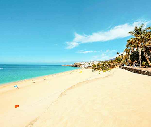 En lang sandstrand med klart blått vann og mennesker som soler og bader. Langs siden er det en promenade med palmer og grøntområder. (AI-generert tekst)