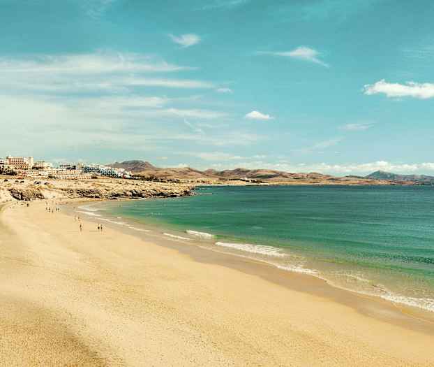En bred sandstrand som strekker seg langs kystlinjen med turkist vann og bygninger i bakgrunnen. (AI-generert tekst)