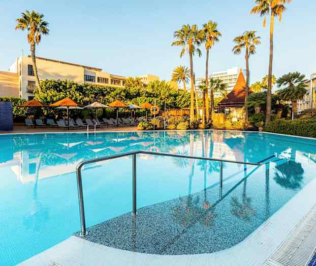 Et utendørs svømmebasseng omgitt av solsenger, parasoller og grønne palmer under en klar blå himmel. (AI-generert tekst)