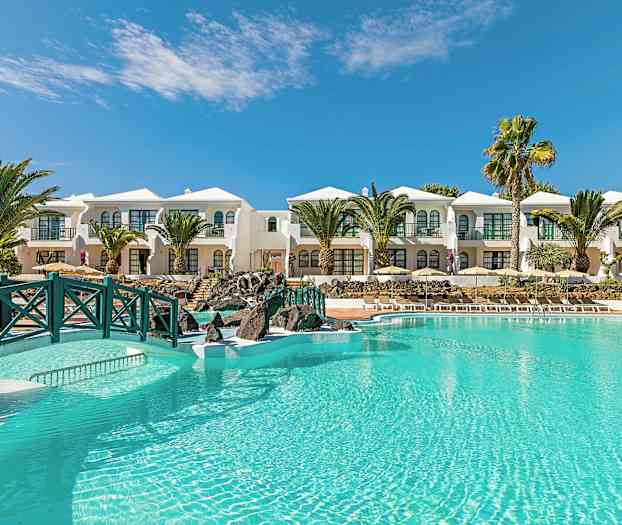 Et lyseblått basseng foran hvite bygninger med palmer og klar himmel i bakgrunnen. (AI-generert tekst)