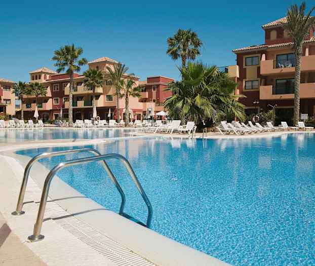 Et feriekompleks med et utendørs basseng omgitt av solsenger og palmer, samt bygninger i bakgrunnen. (AI-generert tekst)