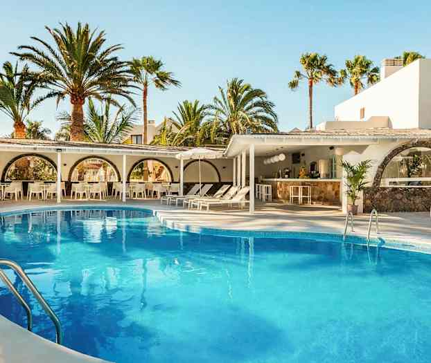 Et utendørs svømmebasseng omgitt av solsenger og parasoller, med hvite bygninger og høye palmer i bakgrunnen. (AI-generert tekst)