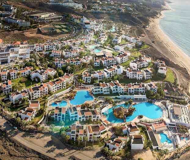 Flyfoto av et feriested med hvite bygninger og røde tak, samt store bassenger og hager nær stranden. (AI-generert tekst)