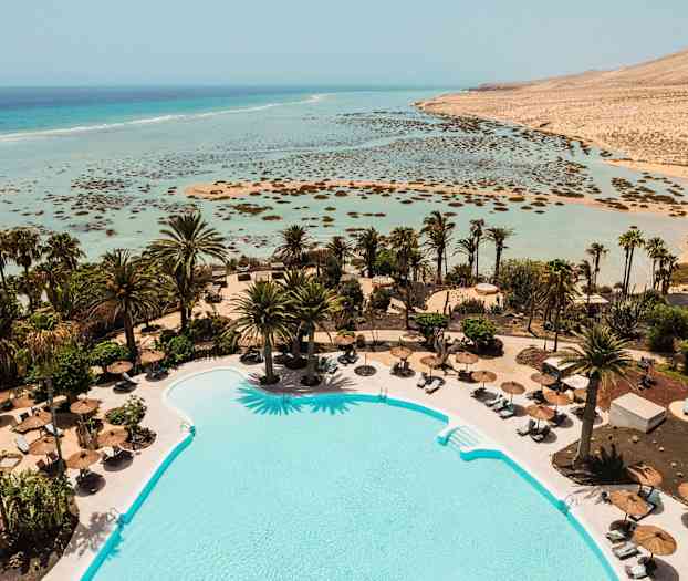 Et basseng omgitt av solsenger og parasoller, med palmer i bakgrunnen og utsikt over en grunne, turkisblå strand og hav. (AI-generert tekst)