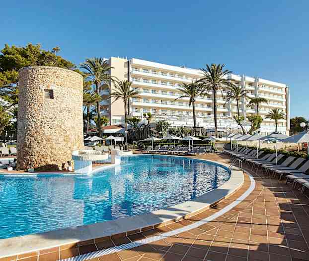 Et svømmebasseng omgitt av solsenger, med et steinbygget tårn og et hotell i bakgrunnen. (AI-generert tekst)