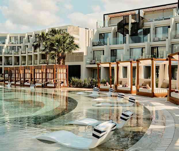 Et hotellbasseng med solsenger i bassenget og cabanas ved siden. Bygningen har balkonger med glassrekkverk. (AI-generert tekst)