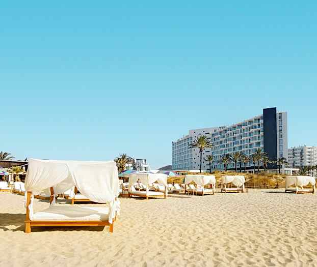 Strand med senger med hvite draperier, i bakgrunnen ses en moderne bygning. (AI-generert tekst)