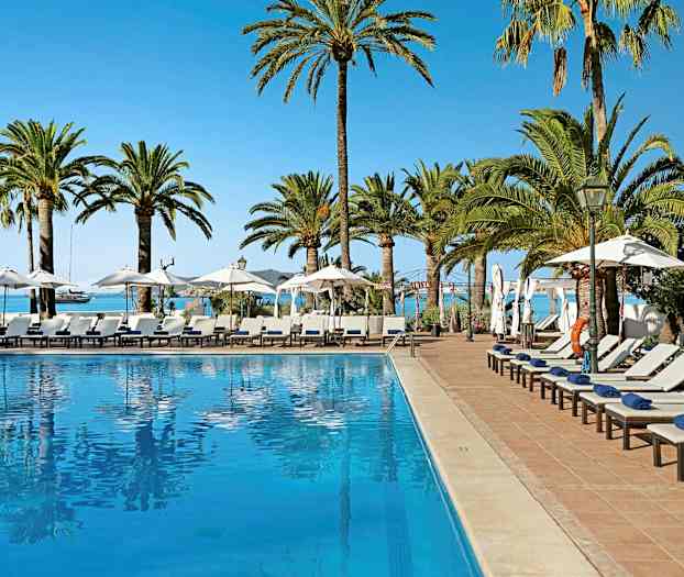 Et utendørs basseng omgitt av solsenger, parasoller og palmer med havet i bakgrunnen. (AI-generert tekst)