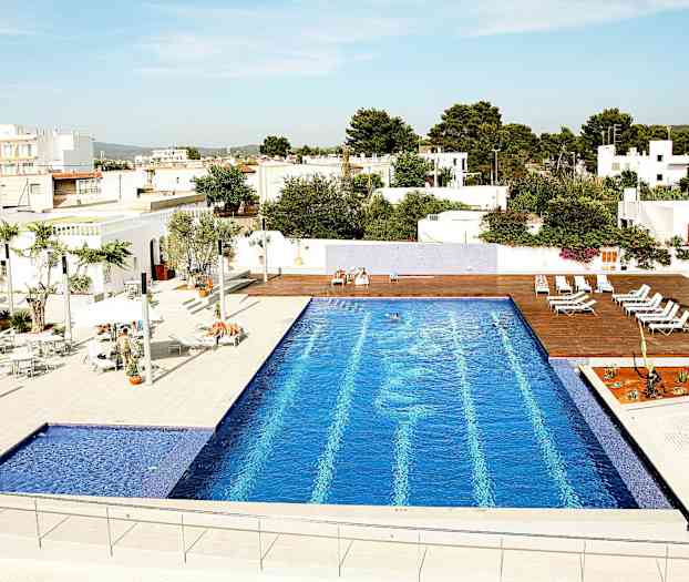 Et utendørsbasseng med blå fliser, omgitt av solsenger og hvite bygninger. (AI-generert tekst)