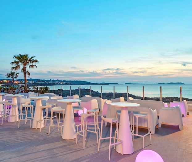 En terrasse med hvite bord og stoler, omgitt av glasspaneler, med utsikt over havet og en palmelignende plante i bakgrunnen. (AI-generert tekst)