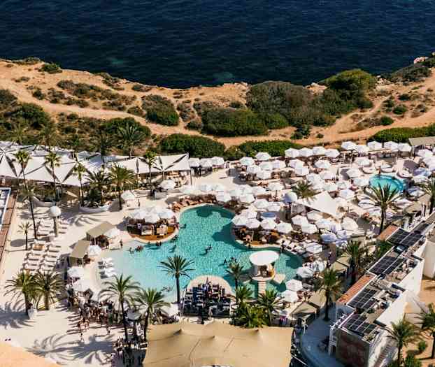 Luftfoto av en ferieanlegg med flere bassenger og hvite parasoller, ved siden av klipper og havet. (AI-generert tekst)