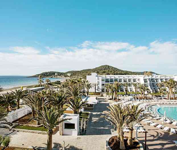 Et hotellområde ved stranden med basseng, palmer og en hvit bygning. (AI-generert tekst)