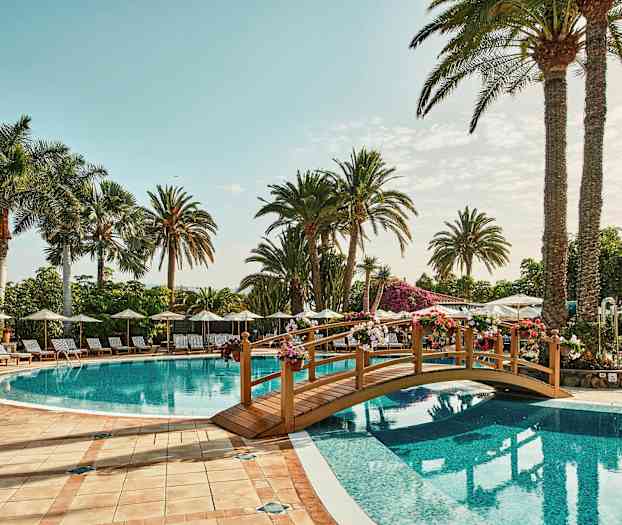 Bilde av et utendørs svømmebasseng med solsenger, parasoller og palmer i bakgrunnen. En liten dekorativ bro spenner over vannet. (AI-generert tekst)