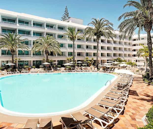 Et hotellbasseng med solsenger og parasoller omgitt av frodige palmer, samt en fleretasjes bygning med balkonger i bakgrunnen. (AI-generert tekst)