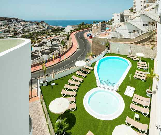 Utendørsbasseng med solsenger på grønn plen og utsikt over nærliggende bygninger og havet. (AI-generert tekst)