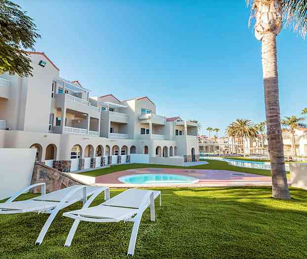 Utendørsområde med solsenger, gressplen, basseng og nærliggende bygning i lyse farger. (AI-generert tekst)