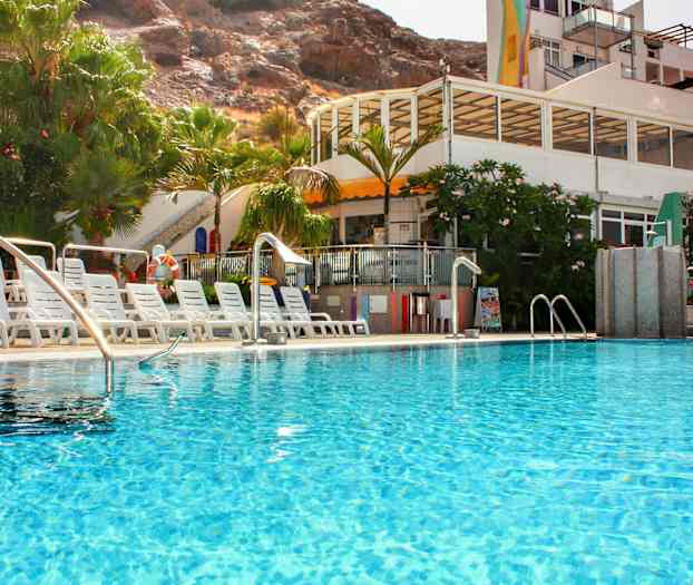 Et utendørs svømmebasseng omgitt av solsenger og grøntområder, med en bygning i bakgrunnen. (AI-generert tekst)