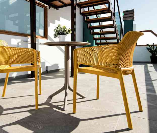 To gule stoler og et bord på en solrik terrasse med trapper og plante. (AI-generert tekst)