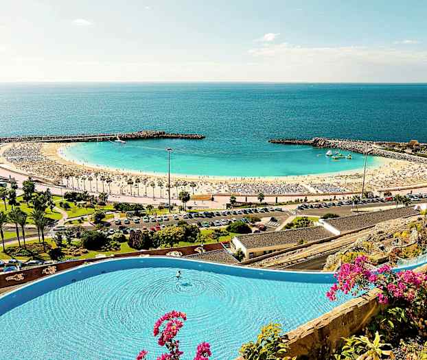 En strand med gul sand og klart blått hav, omgitt av en beskyttende molo og grøntområder. Et basseng med blomster i forgrunnen. (AI-generert tekst)