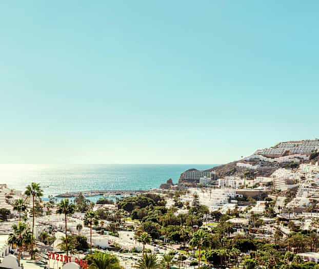 Utsikt over en kystby med hvite bygninger og palmer foran et glitrende hav. (AI-generert tekst)